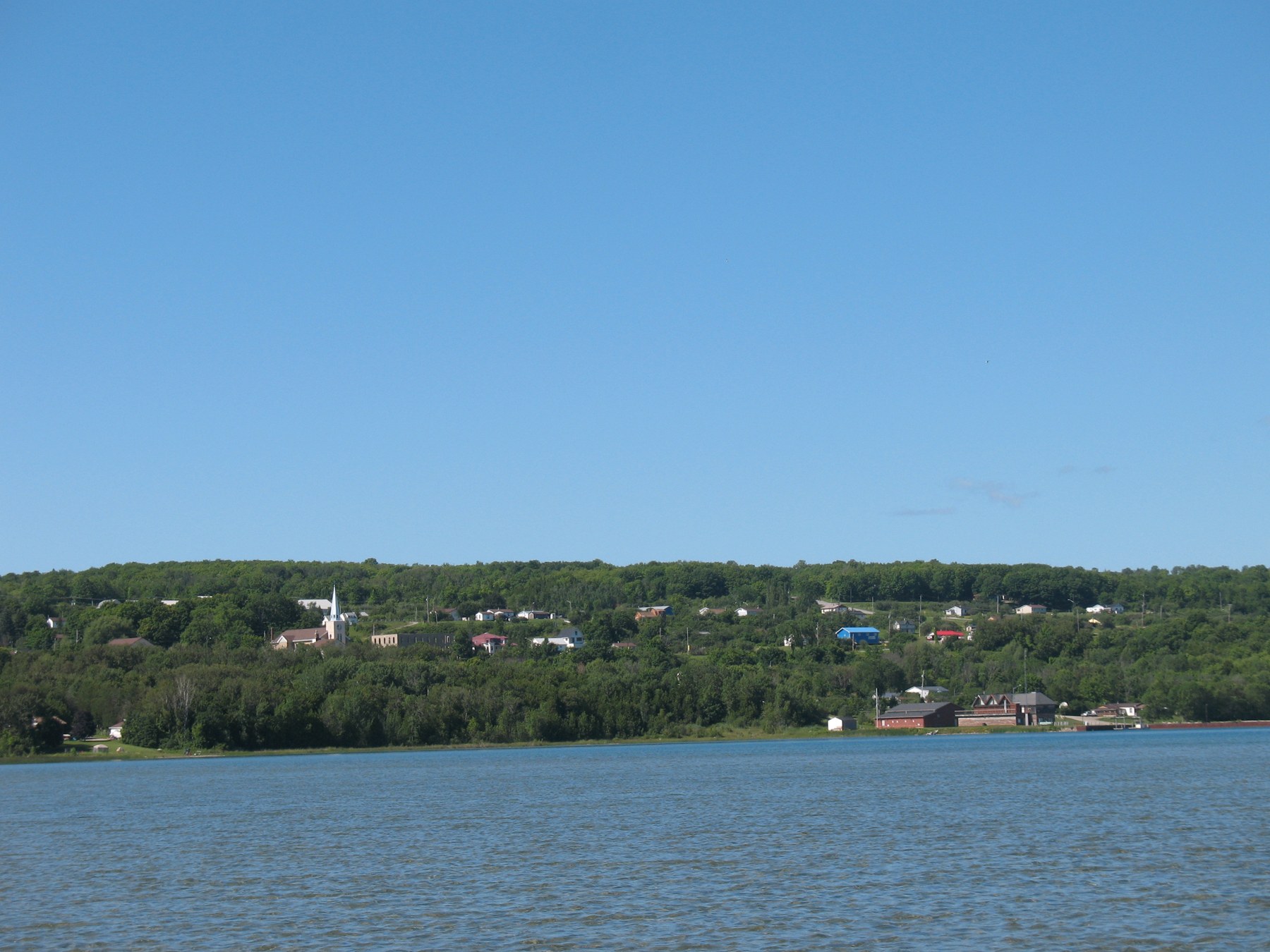 View across blue water toward wooded Wiikwemkoong, with buildings and a church steeple along the shore.