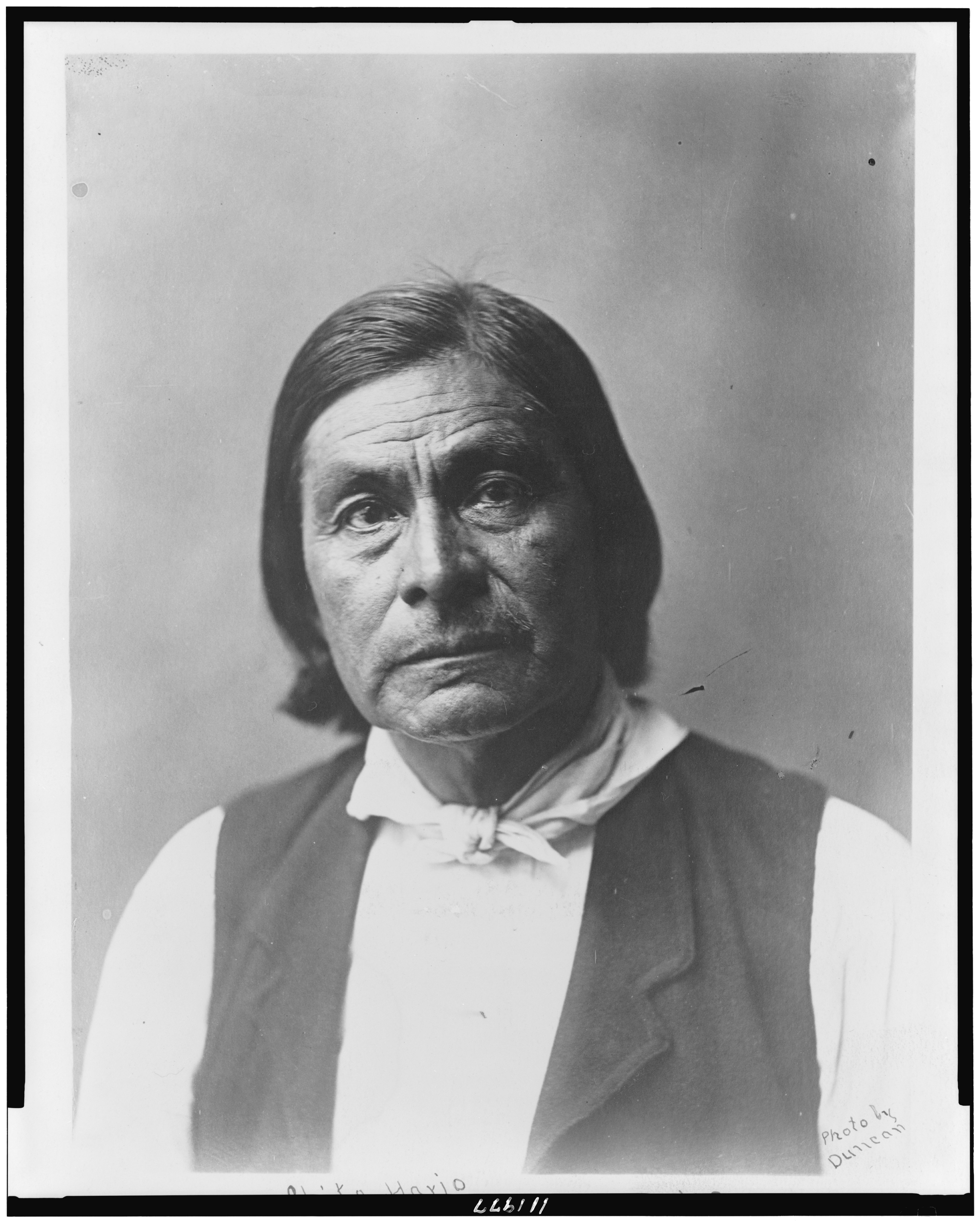 Black-and-white head-and-shoulders portrait of Chitto Harjo facing forward, with dark shoulder-length hair, a light shirt and a dark vest; the photographic copy includes print margins and Duncan's credit.