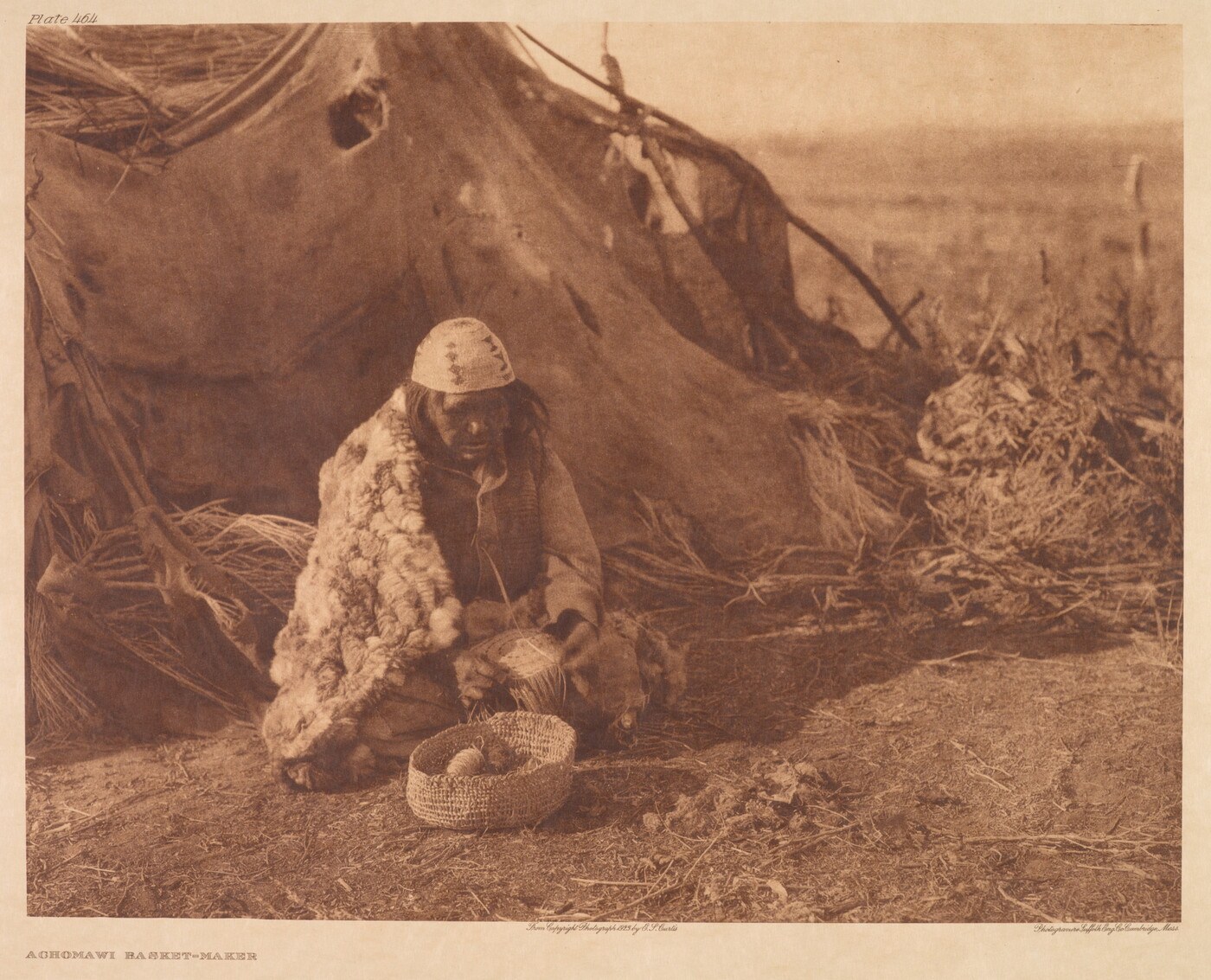 Sepia photogravure of an unnamed Achomawi basketmaker seated with a basket in progress and a finished basket in front of a shelter.