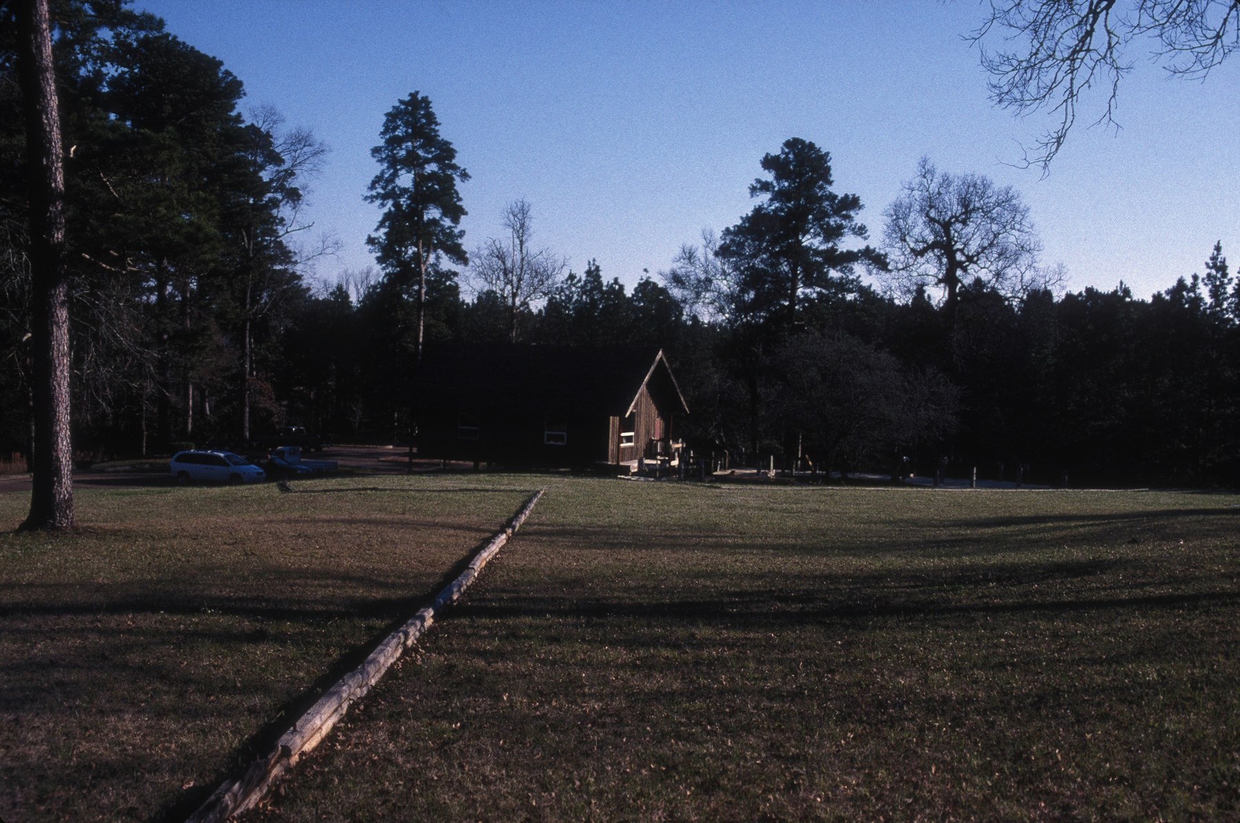 A grassy clearing and marked presidio outline at Los Adaes State Historic Site, with pine trees, a modern visitor building and parked vehicles beyond.