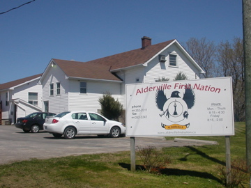 A white administration building beside a public sign reading Alderville First Nation, with parked cars and a grassy foreground.