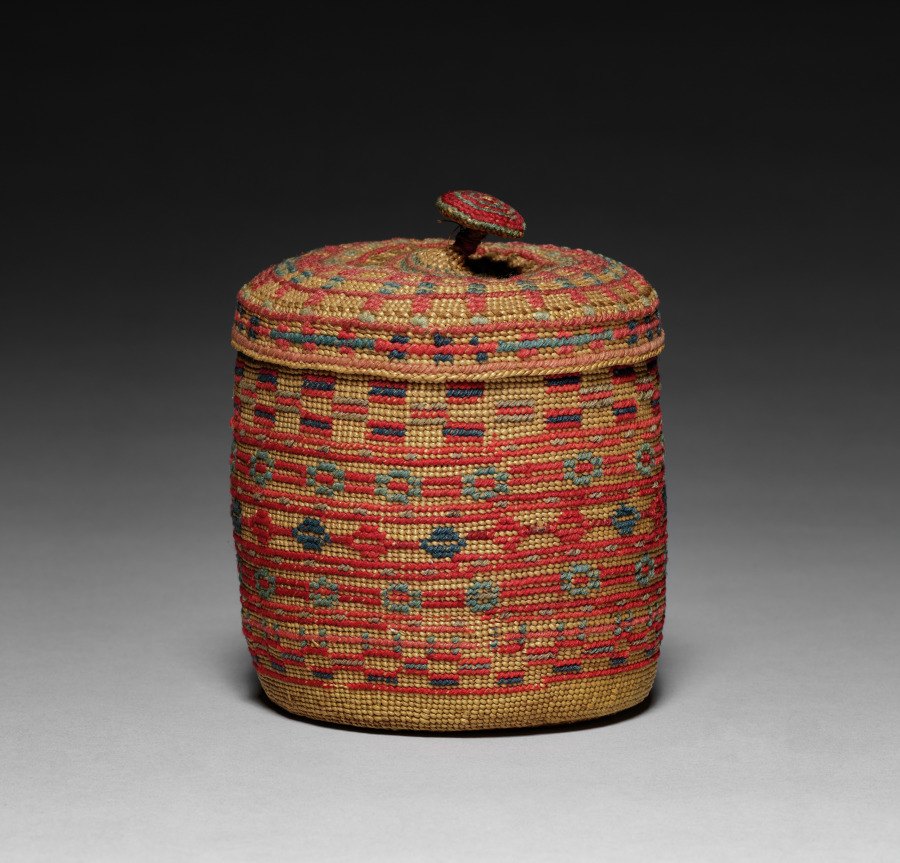 Full frame shows a cylindrical covered basket with red, tan, green and blue wool decoration against neutral gray.