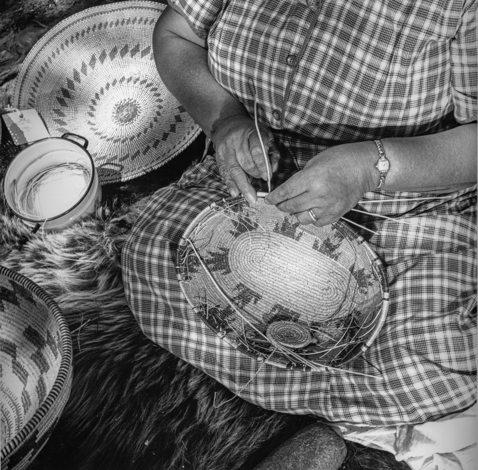 Black-and-white close-up of Selena LaMarr’s hands weaving a coiled basket, with completed baskets and materials beside her.