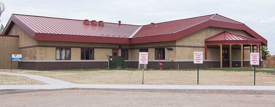 A low college building with a red metal roof, tan exterior walls and roadside signs.