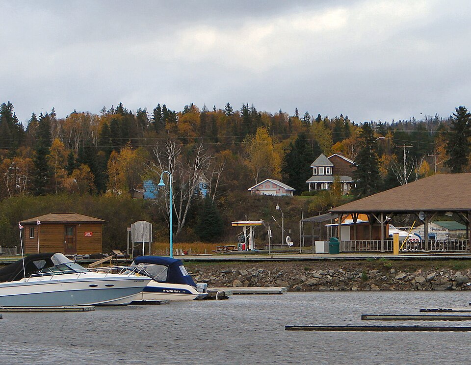 A waterfront village scene with boats and dock, small buildings and autumn trees.