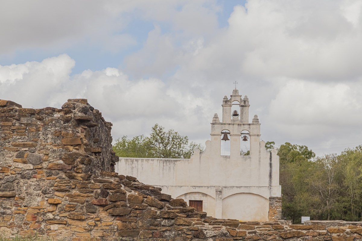 The white church and bell wall at Mission San Juan stand behind low stone ruins beneath a cloudy sky.