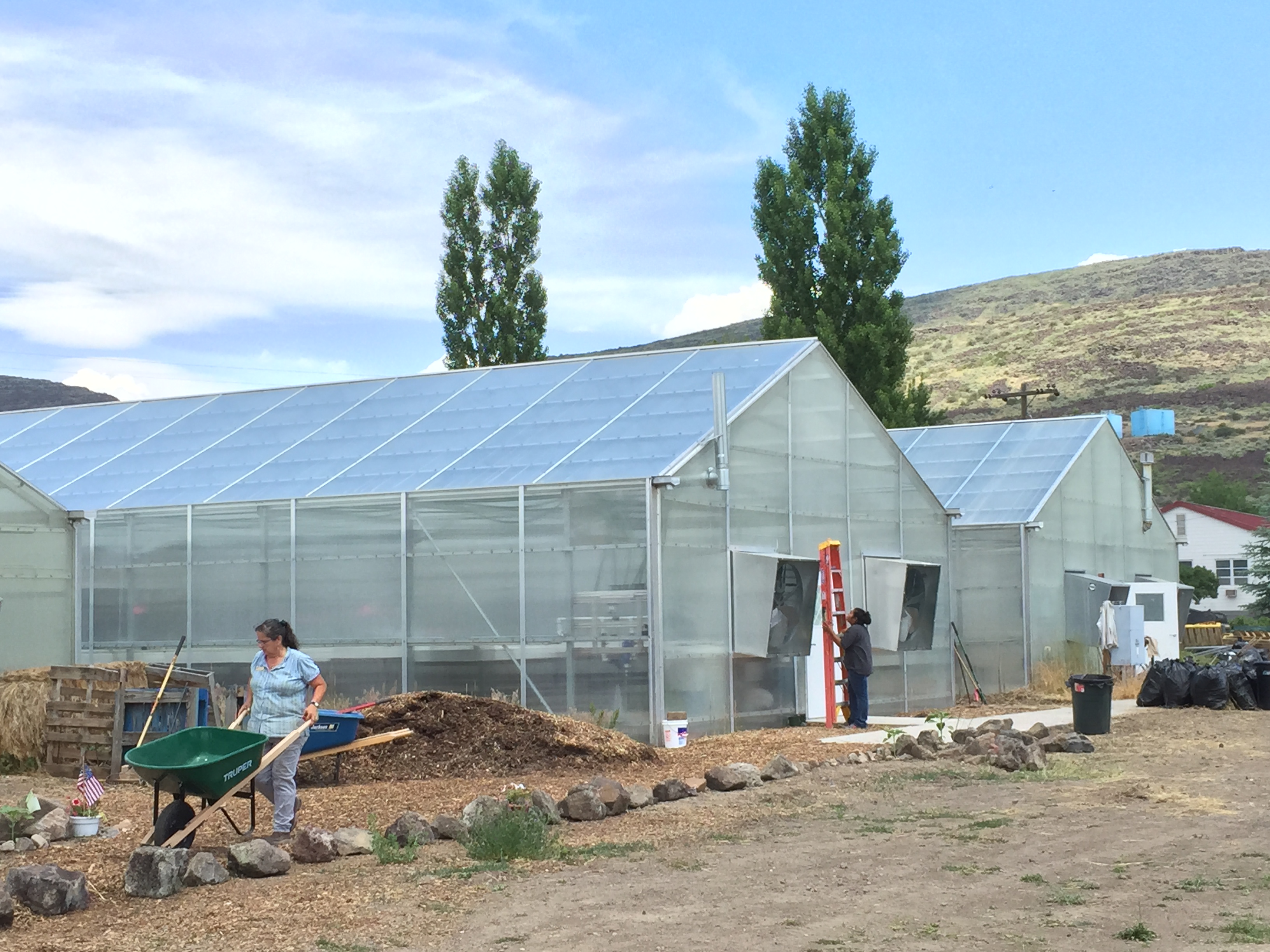 Greenhouses at Duck Valley with two people working outside, a wheelbarrow, and dry hills behind.