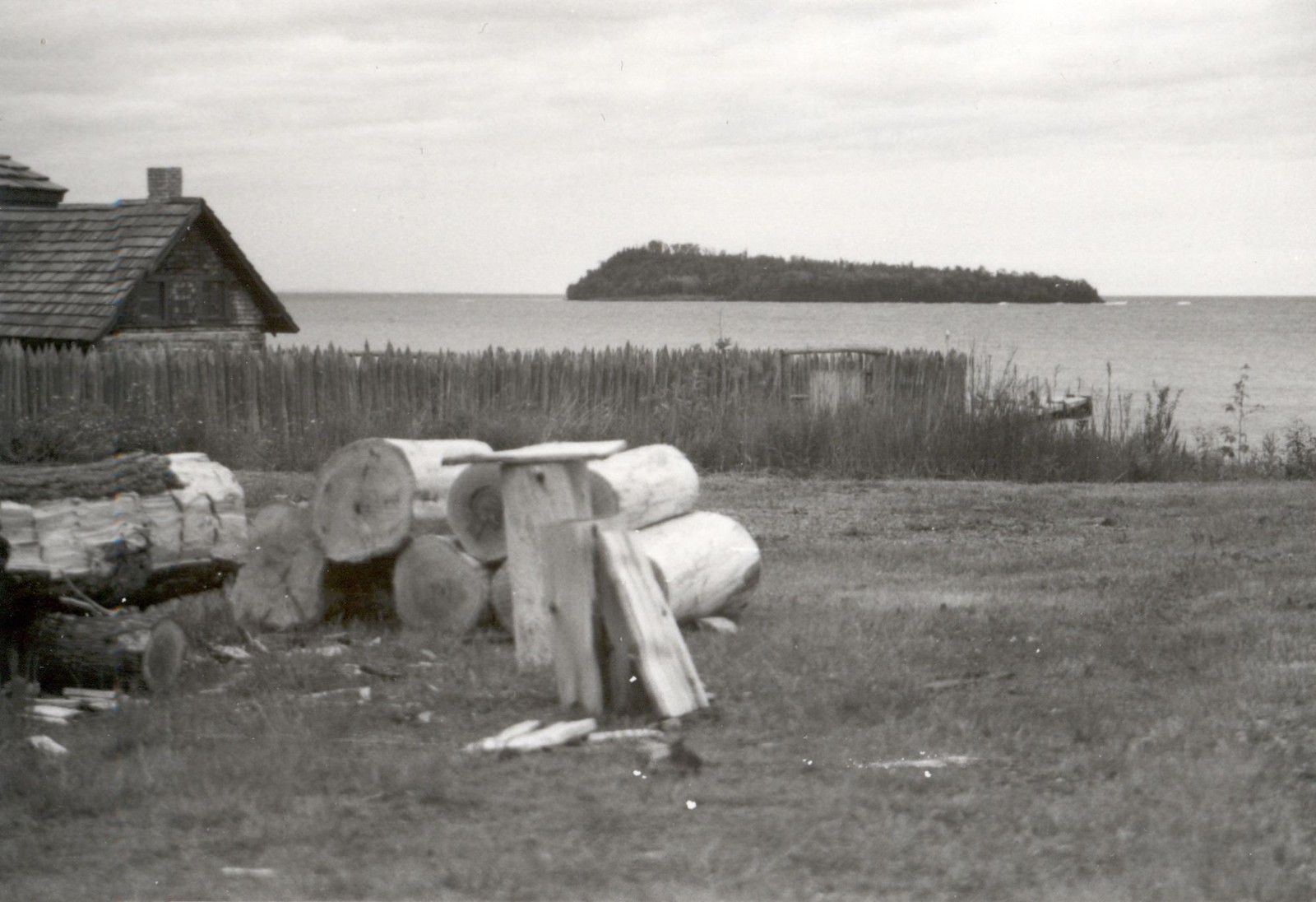 Black-and-white 1984 view of Grand Portage Bay and a wooded island beyond the reconstructed depot’s log structures and stockade.