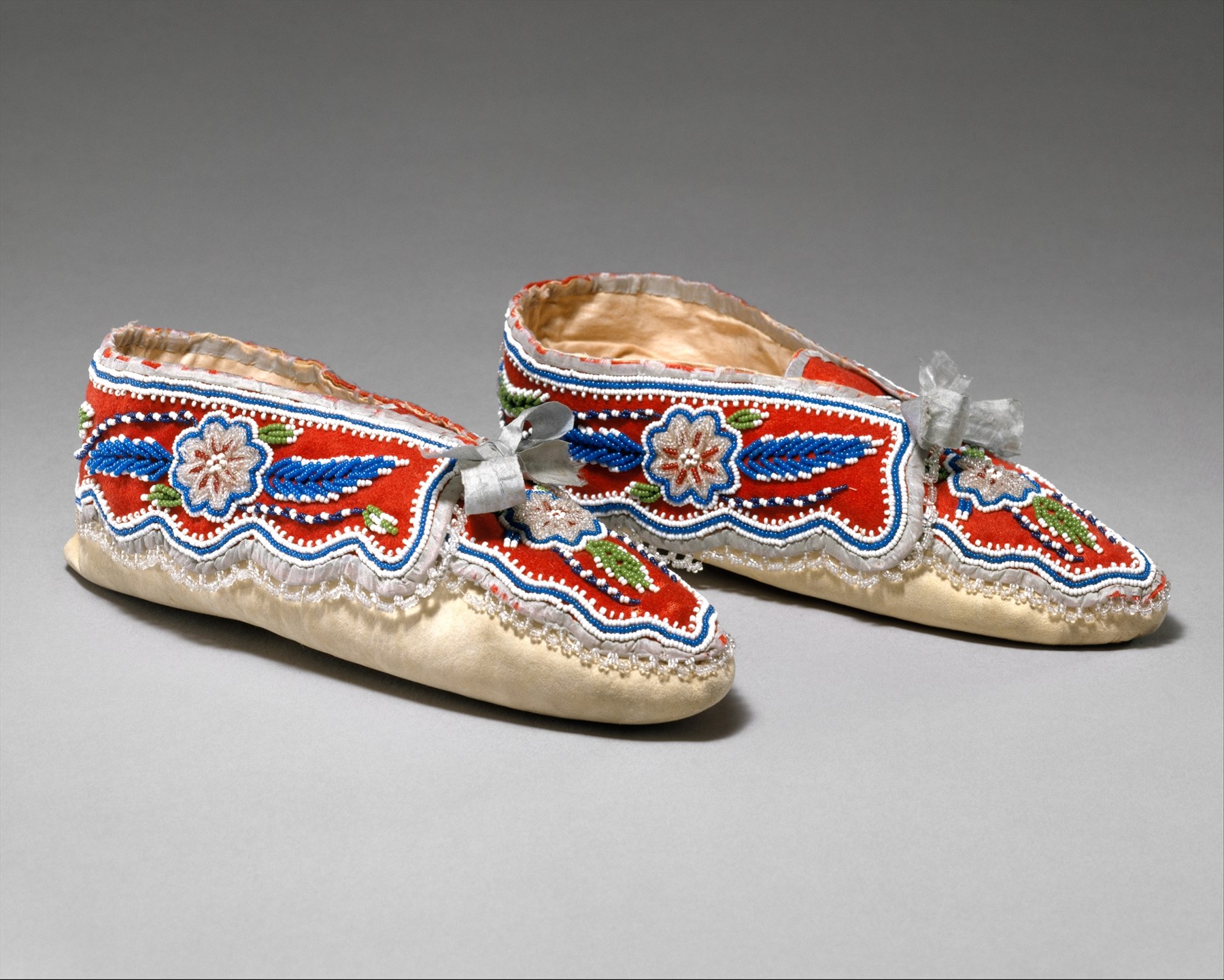 A pair of red and cream moccasins decorated with white, blue and green beadwork, photographed on gray.