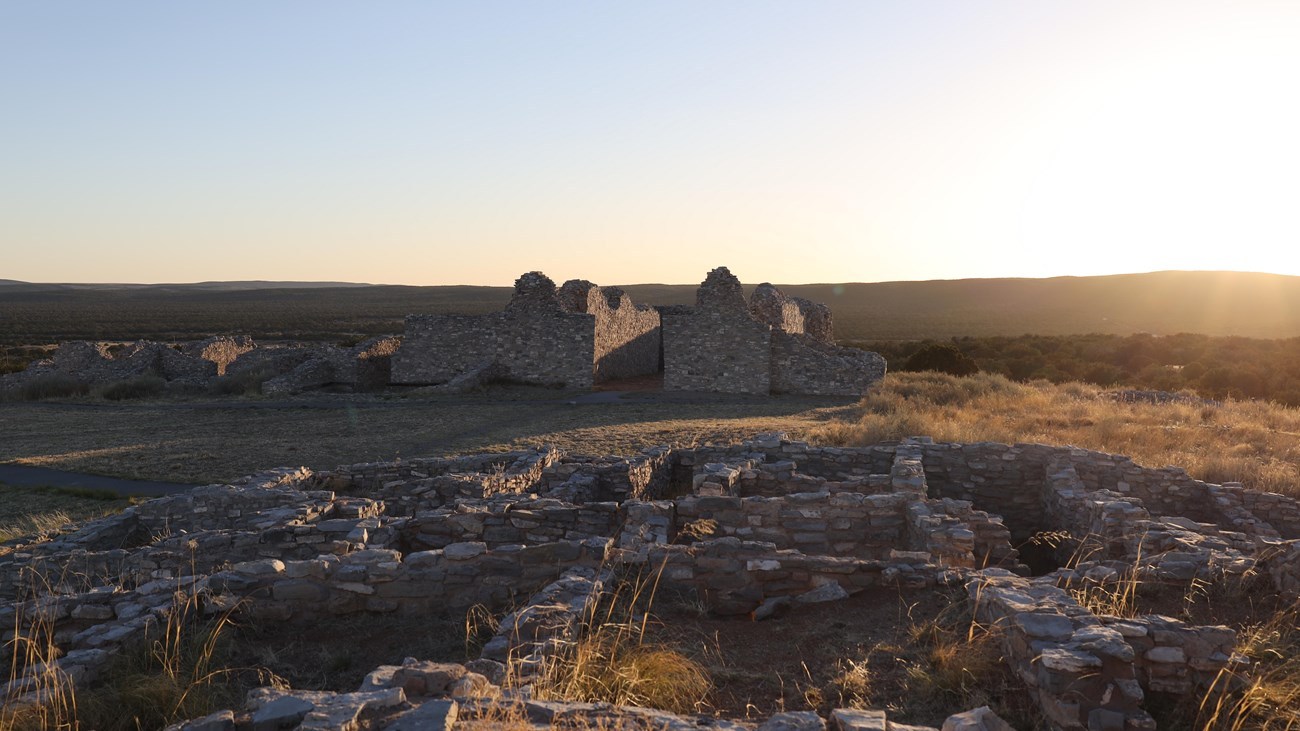Low limestone pueblo remains at Gran Quivira lie in the foreground, with roofless church walls and open countryside in warm low sunlight.
