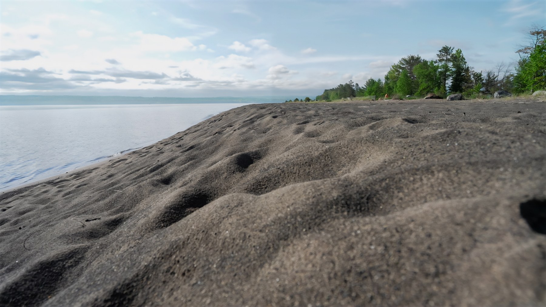 Dark stamp sands beside calm water, with trees along the shore in a Forest Service photograph discussing Keweenaw Bay Indian Community restoration.