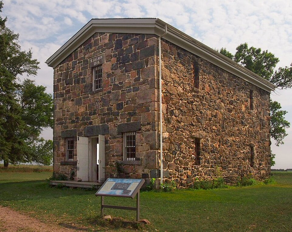 A stone warehouse with a pale roof and open door, lawn, trees and an interpretive sign at the Lower Sioux Agency historic site.