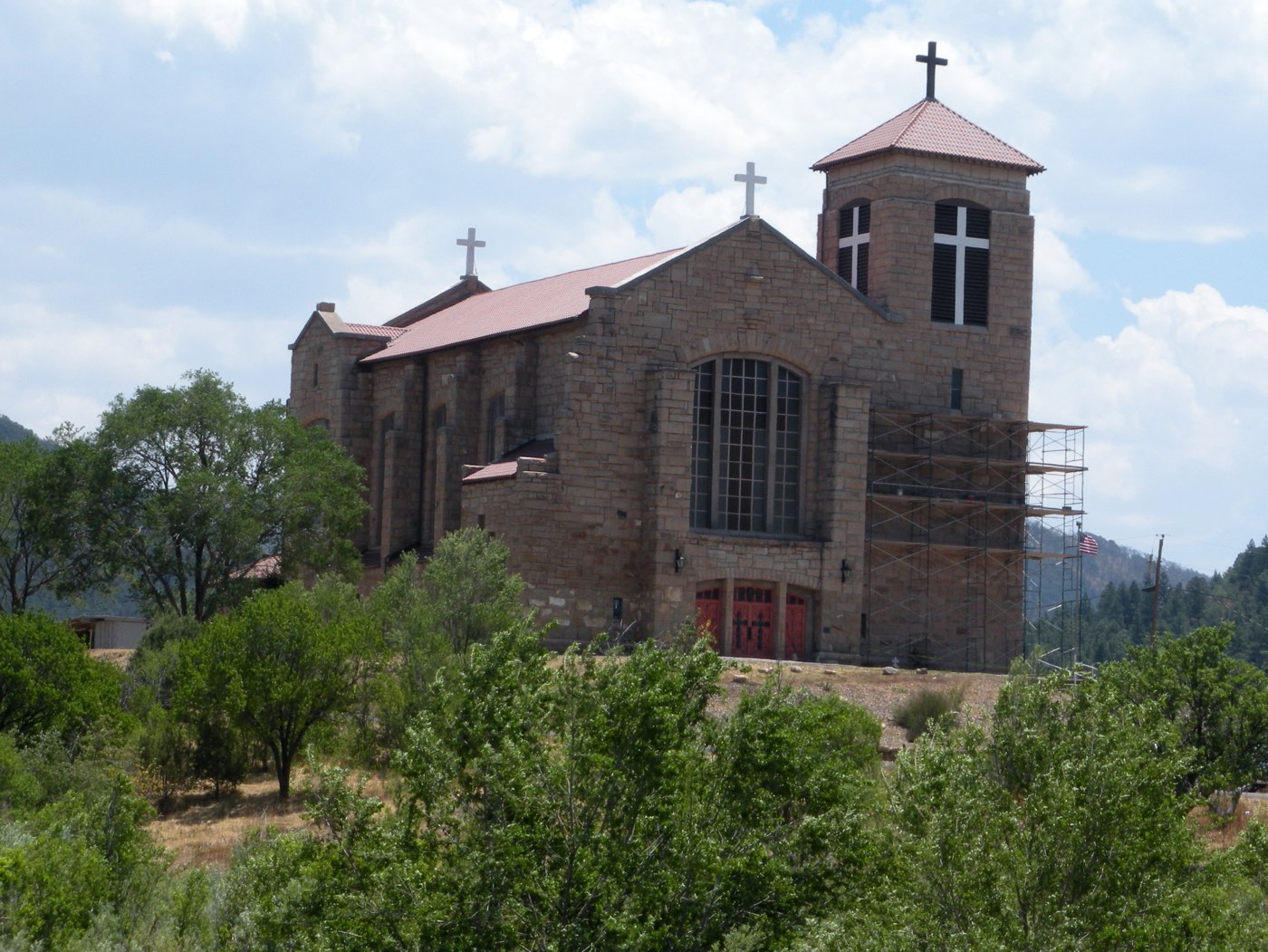 Stone exterior of St. Joseph Apache Mission in Mescalero, with a square bell tower, red doors, restoration scaffolding, and trees in the foreground.