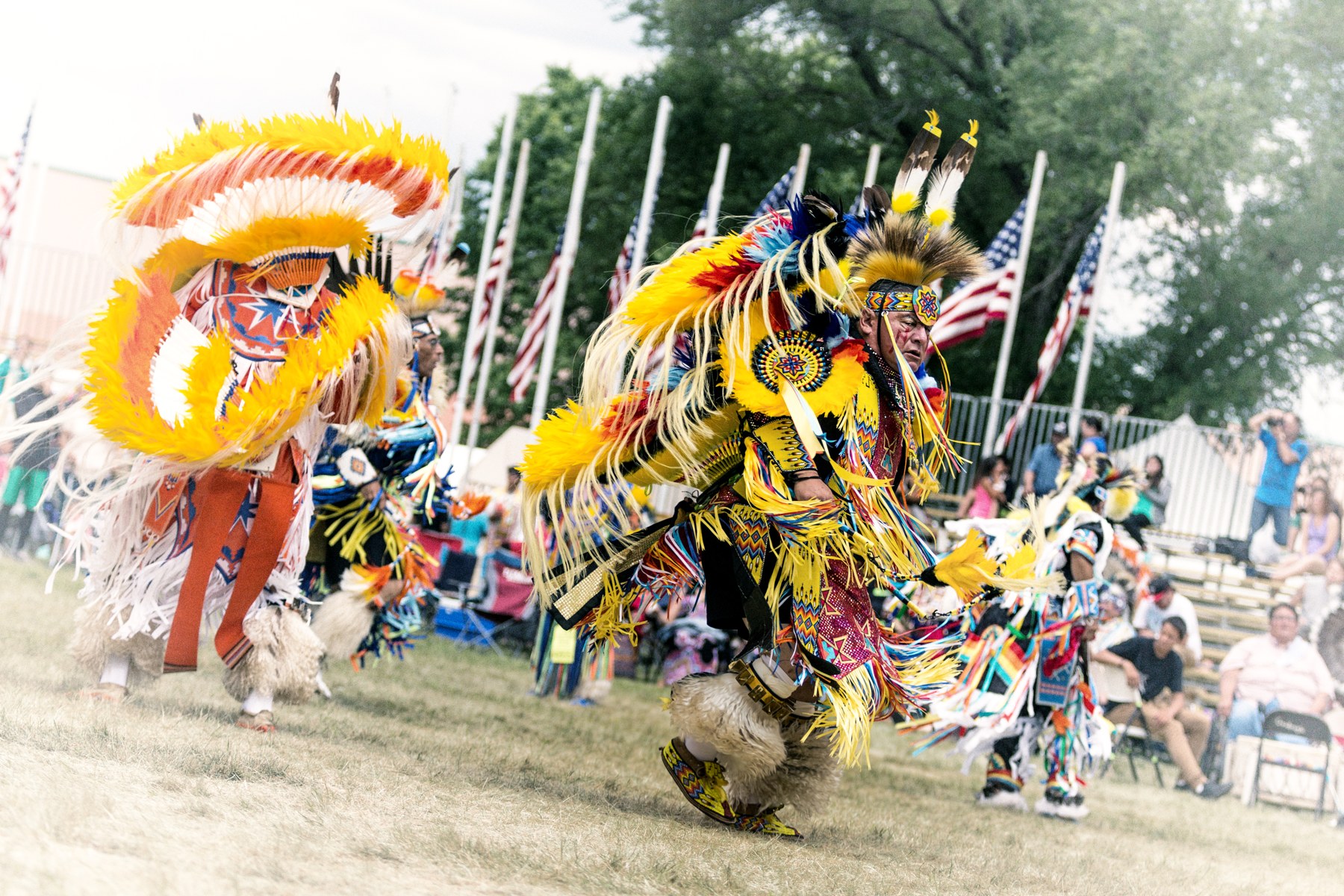 Dancers in colorful regalia at the 2017 Prairie Island Indian Community Wacipi, with spectators and flags behind them.