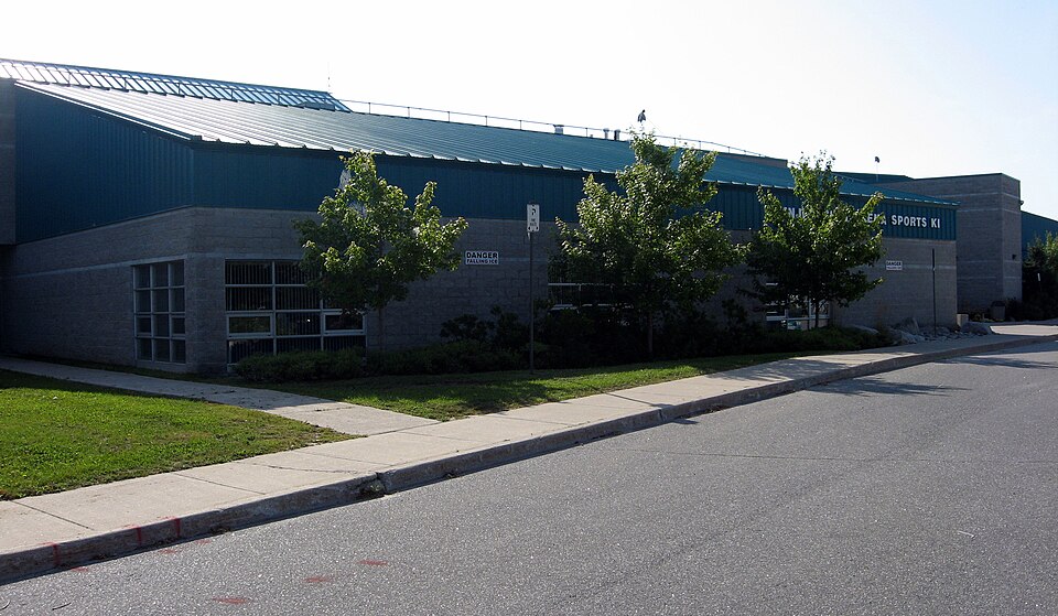 Exterior of Mnjikaning Arena Sports Ki at Rama, with trees, a sidewalk and road in front of the building.