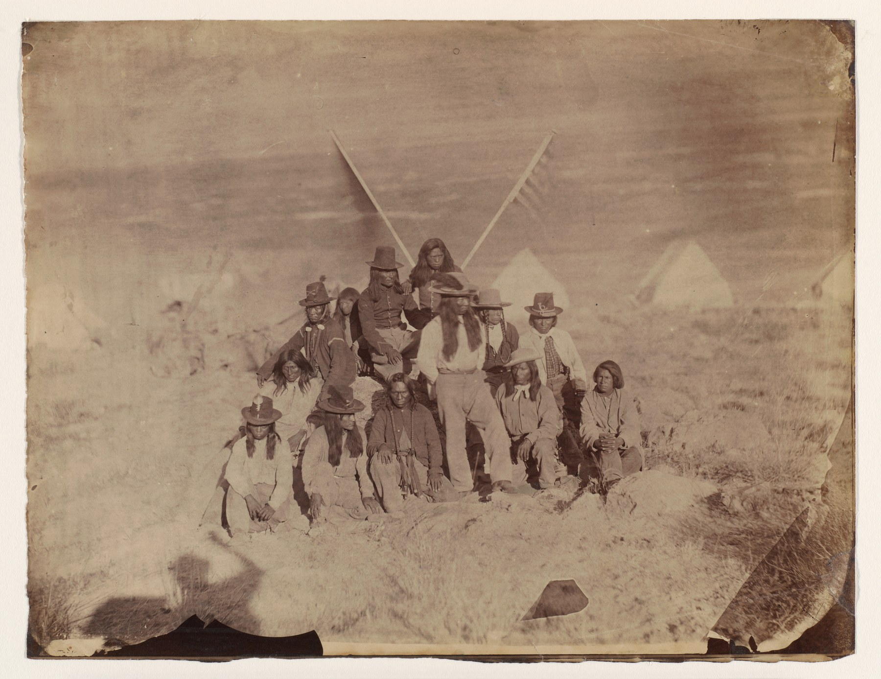 Sepia archival group portrait catalogued as Shoshone, with tents and crossed flagpoles behind the group; the complete print has worn edges.