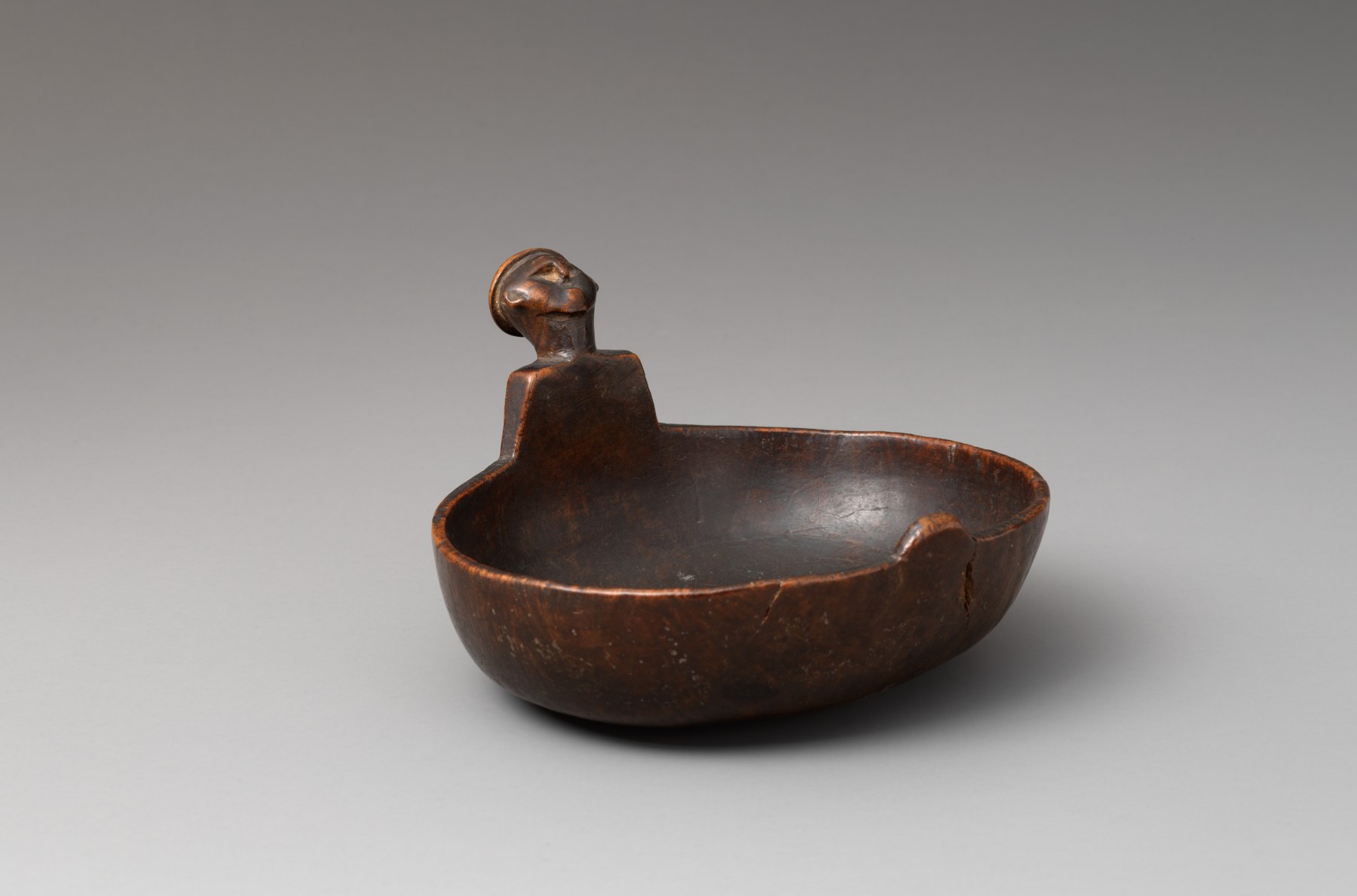 A dark wooden bowl with an upright carved human figure at its rim, photographed on gray.