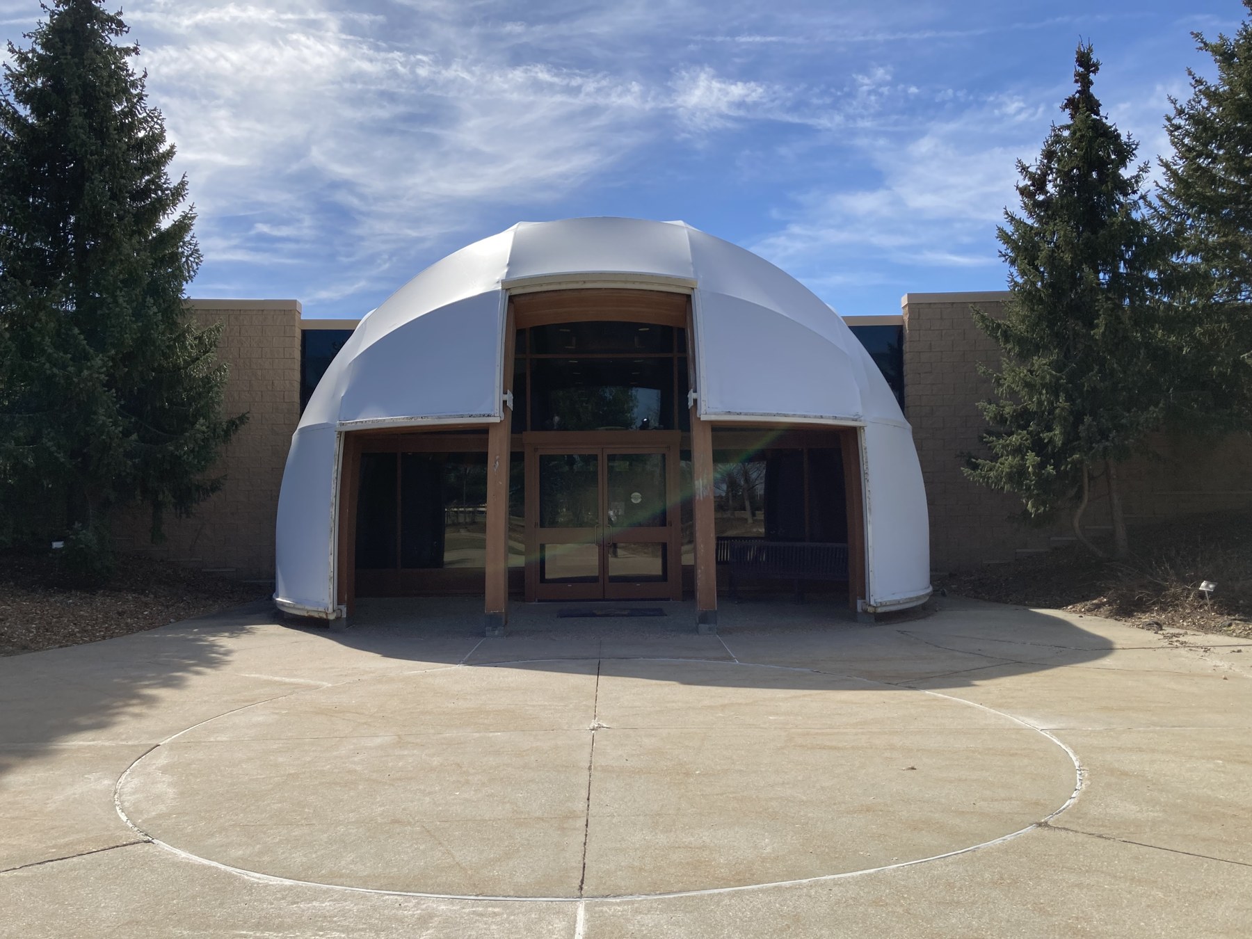 Curving white entrance of the Ziibiwing Center, with trees, pavement and a partly cloudy sky.